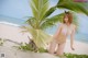 A woman in a bikini sitting under a palm tree on the beach.