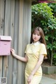 A woman in a yellow dress leaning against a wooden wall.